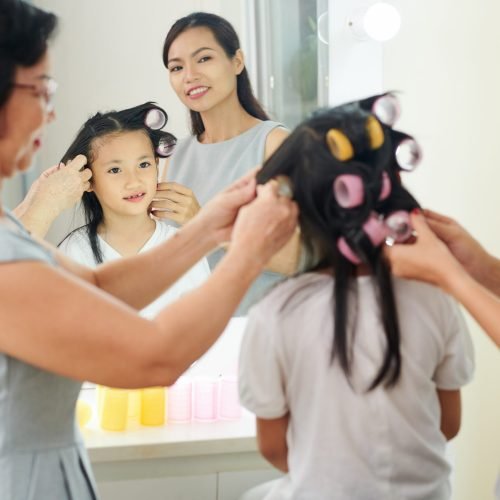 Mother and grandmother curling curlers to little girl together in front of the mirror and preparing for holiday event